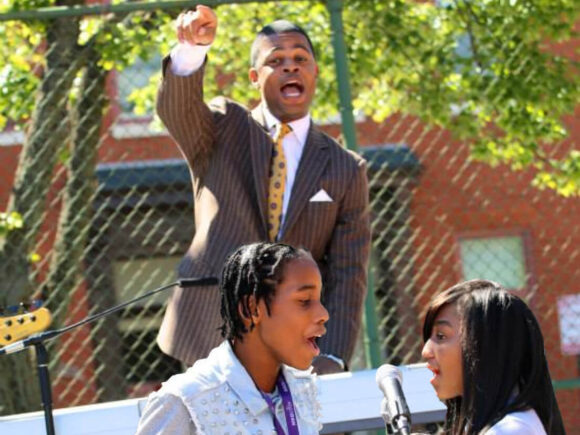 Three people performing outdoors: two singing at microphones in the foreground, one in the background pointing and appearing to lead the performance.