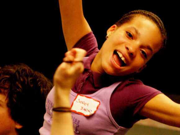 A person wearing a purple shirt with a name tag smiles widely, raising their arms.